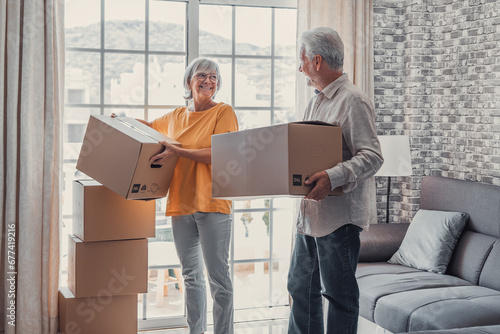 Foto Mature couple moving into new apartment, carrying cardboard boxes into empty room with potted plants
