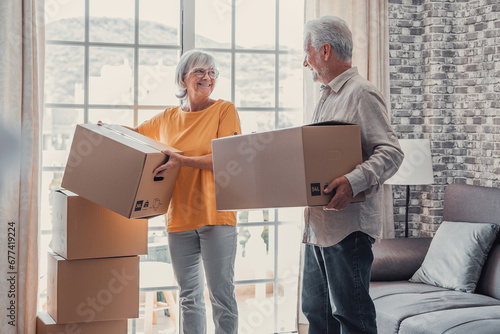Mature couple moving into new apartment, carrying cardboard boxes into empty room with potted plants. Real estate property buying, relocation, new home concept. Rear view. © Daniel
