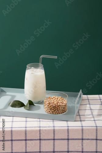 A tray in pastel color featured a milk cup with straw and a transparent bowl of soybeans. Green background. Soy milk is a well-known dairy milk replacement
