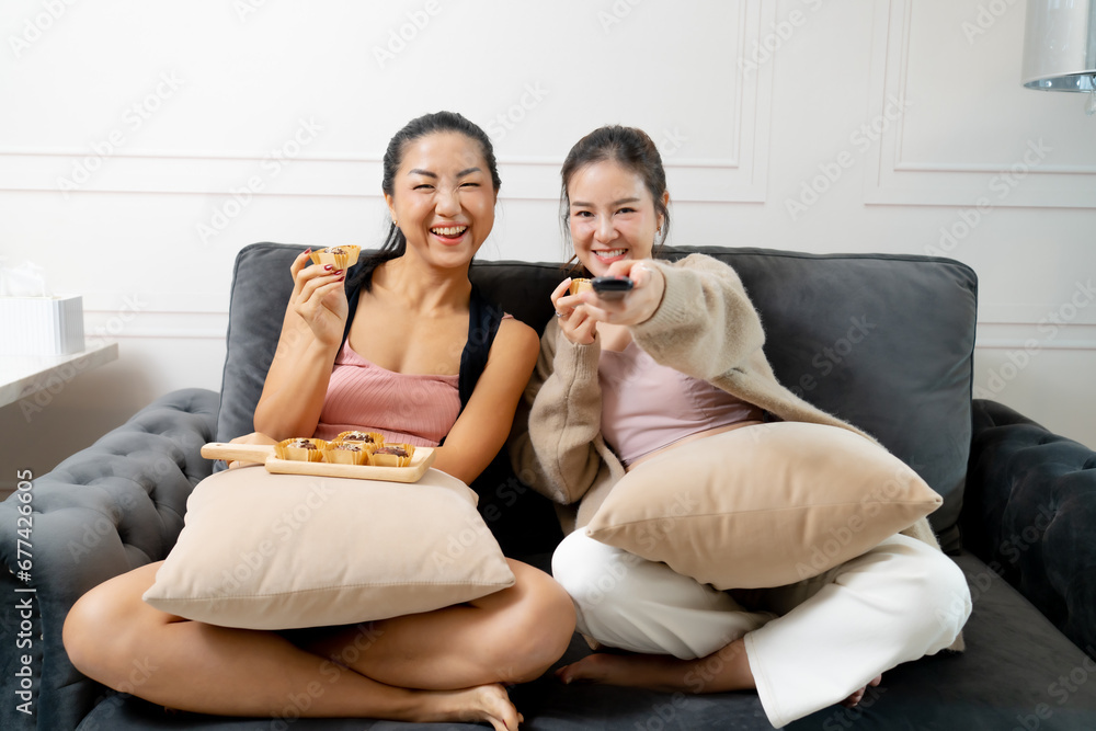 Young Asian woman sitting Sit and relax with friends and eat snacks on the couch, she is using the remote control and choosing a TV show or movie on the television menu in her house on holidays.