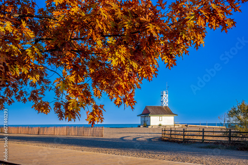 fall color: bright orange maple leaves in autumn with a wooden board walk and snow fence in foreground kew beach, the iconic leuty lifeguard station and lake ontario in the background with blue sky