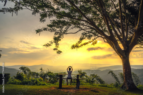 A man sits on a wooden table watching the sunset and use phone to shoot next to a big tree. Travel, vacation concept.