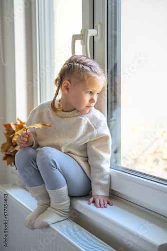 Little sad girl in warm sweater and socks sitting by window, holding autumn leaf and warming up from the heating radiator. Heating in an apartment, at home.