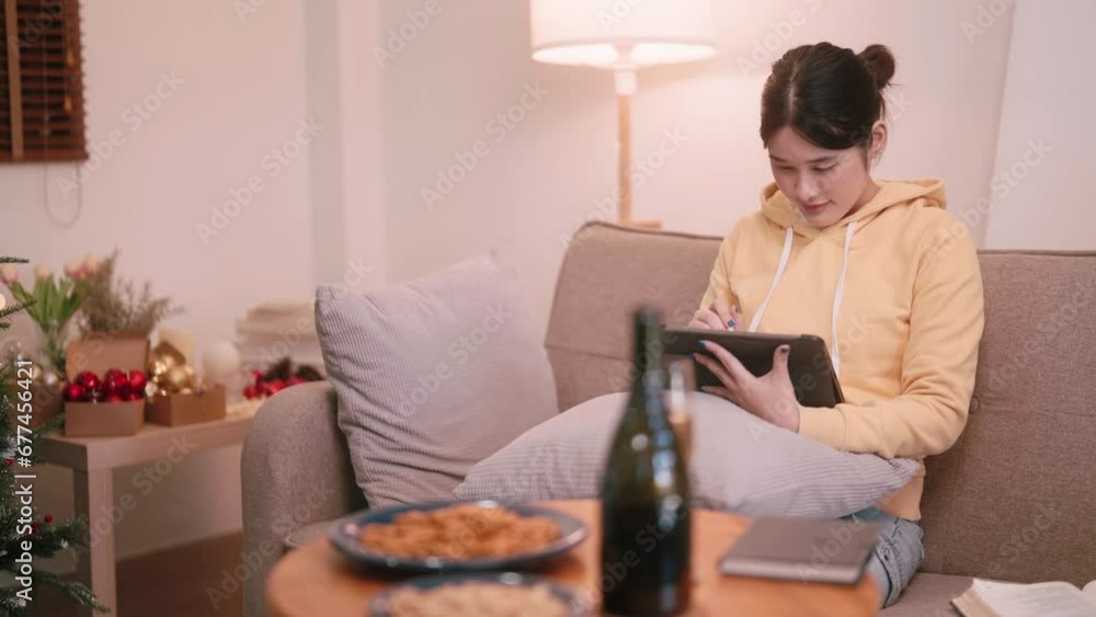 Asian young woman unwinds with tablet drawing on the sofa in the cozy living room during the Christmas evening. There are snacks and drinks on a nearby table for a creative and delightful holiday. Asian young woman unwinds with tablet drawing on the sofa in the cozy living room during the Christmas evening. There are snacks and drinks on a nearby table for a creative and delightful holiday.
