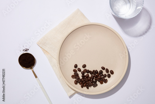 Against the white background, coffee beans and coffee powder contained on ceramic dish and spoon, decorated with glass cup and beige fabric. Blank space on dish for product presentation