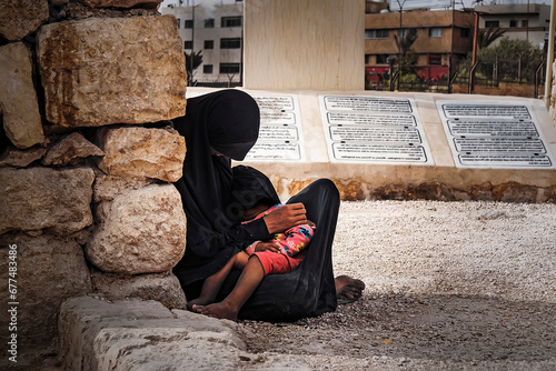 poor Muslim woman in a black hijab sits on the ground and holds her baby in her arms against the backdrop of a city in the Middle East. concept of poverty and misery. Internally displaced persons.