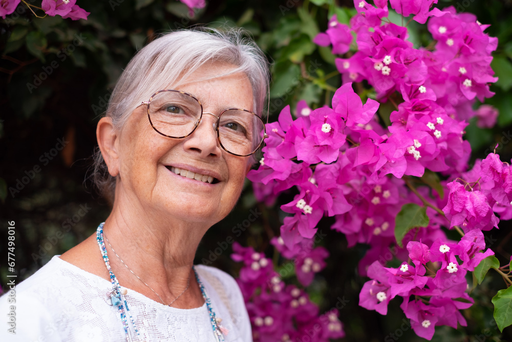 Outdoor close-up portrait of a smiling elderly woman near a pink bougainvillea flower plant in the park. Aged white-haired pensioner in a serene expression loves nature Outdoor close-up portrait of a smiling elderly woman near a pink bougainvillea flower plant in the park. Aged white-haired pensioner in a serene expression loves nature