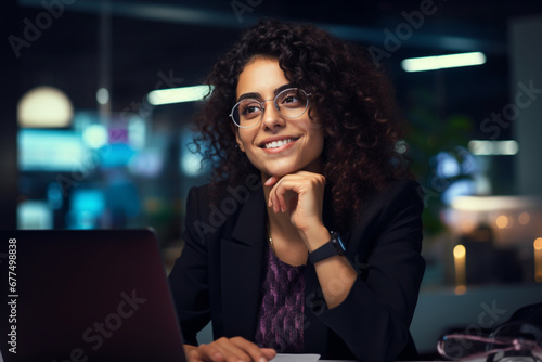 Portrait of a Happy Middle Eastern Manager Sitting at a Desk in Creative Office, Stylish Female with Curly Hair Using Laptop Computer, Thinking About a Business Strategy in Marketing Agency at Night