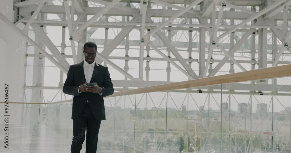Cheerful African American businessman walking in office center checking smartphone and smiling. Beautiful urban landscape visible in panoramic window. Cheerful African American businessman walking in office center checking smartphone and smiling. Beautiful urban landscape visible in panoramic window.
