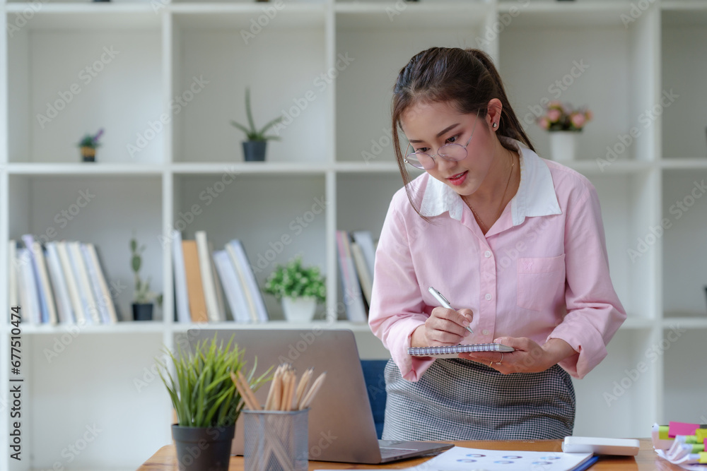 Young Asian businesswoman using laptop computer, taking notes, working project in modern office. Financier working with documents at workplace. Young Asian businesswoman using laptop computer, taking notes, working project in modern office. Financier working with documents at workplace.