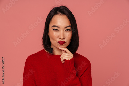 Pensive asian woman touching her chin and looking at camera while standing isolated over pink wall