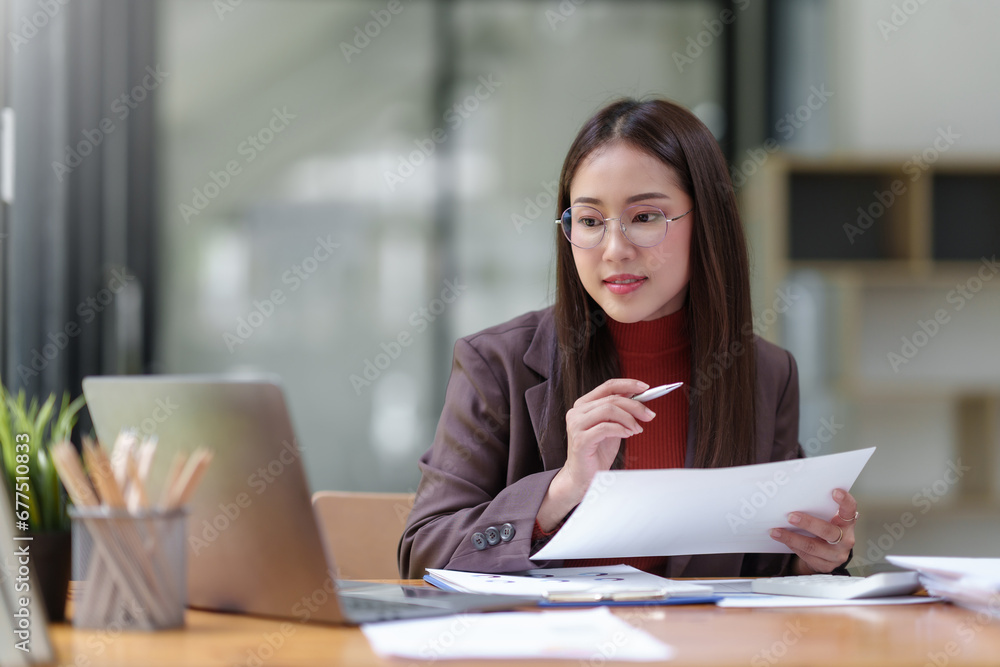 Young Asian businesswoman using laptop computer, taking notes, working project in modern office. Financier working with documents at workplace. Young Asian businesswoman using laptop computer, taking notes, working project in modern office. Financier working with documents at workplace.