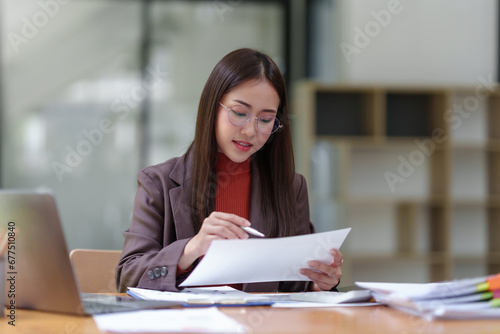 Young Asian businesswoman using laptop computer, taking notes, working project in modern office. Financier working with documents at workplace.