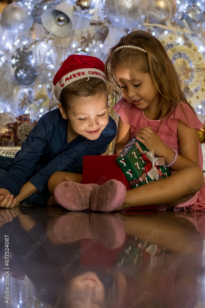 Obraz premium Two excited little children siblings- brother and sister opening a magical gift box by cozy Christmas tree. Merry Christmas and happy holidays
