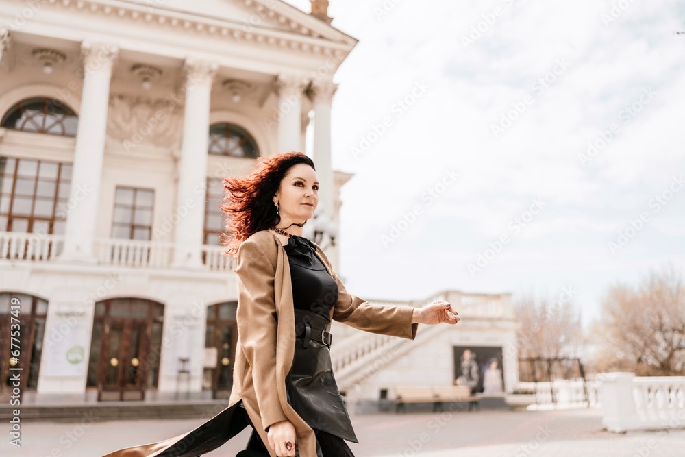 Woman street lifestyle. Image of stylish woman walking through European city on sunny day. Pretty woman with dark flowing hair, dressed in a beige raincoat and black, walks along the building.