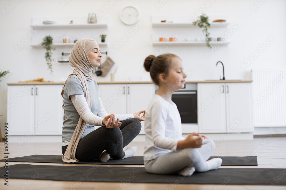 Fototapeta premium Side view focus on peaceful young woman in hijab meditating in seated asana with adorable girl indoors. Calm mother and little daughter working out on yoga mats together on sunny day at home.