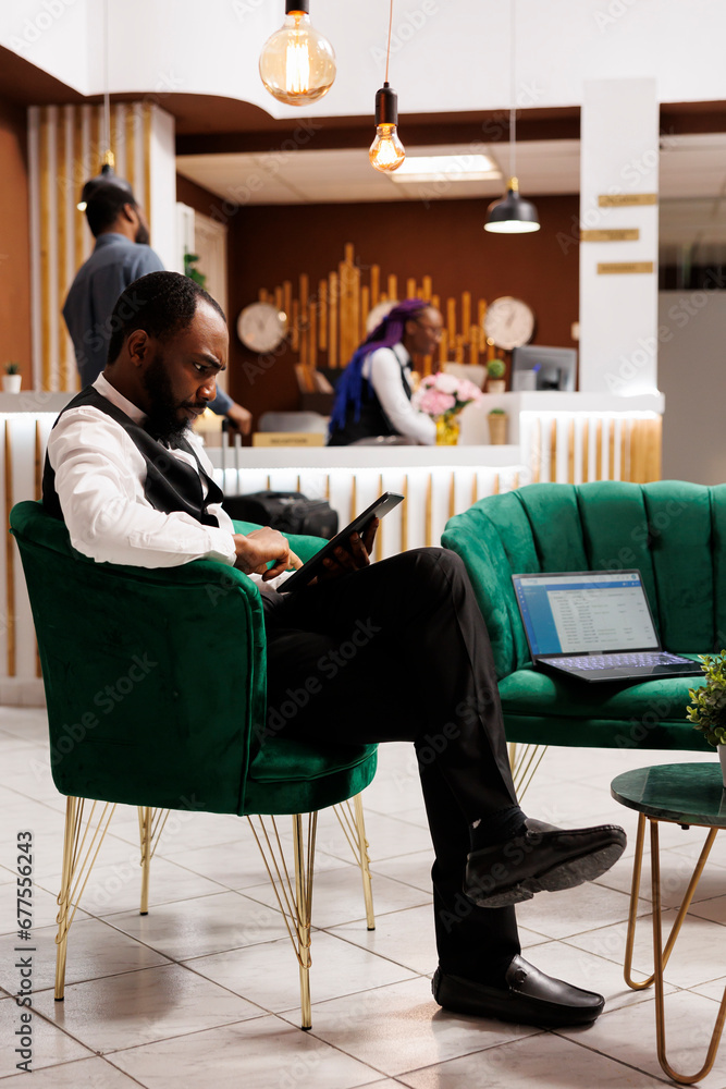 Focused concentrated hotel manager holding digital tablet using front desk software, confirming online bookings while sitting in lobby. Innovative technologies in hospitality industry
