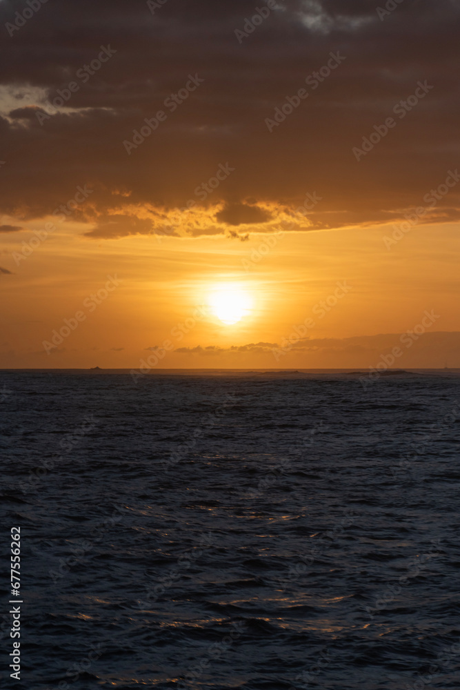 A dramatic orange sunset with colorful clouds and dark blue water from a boat on the Pacific Ocean off of the Island of Kauai in Hawaii, United States.
