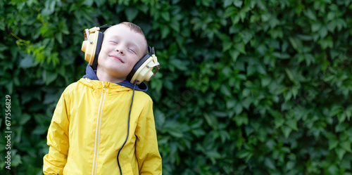A positive little boy of 6 years old in a yellow jacket and yellow retro earphones listens to music. The boy stands against the background of a wall of plants. Banner with space for text