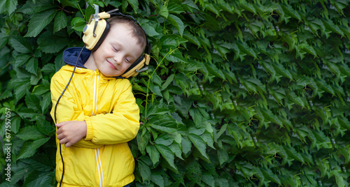 A positive little boy of 6 years old in a yellow jacket and yellow retro earphones listens to music. The boy stands against the background of a wall of plants. Banner with space for text