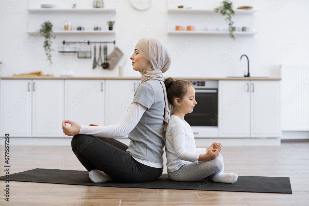 Energetic little girl sitting back to back with muslim lady during her achieving cross-legged position. Happy mother promoting daughter's sense of calmness by meditating together at home. Energetic little girl sitting back to back with muslim lady during her achieving cross-legged position. Happy mother promoting daughter's sense of calmness by meditating together at home.