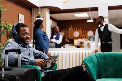 Happy African American guy hotel guest holding digital device making video call while relaxing in lobby. Relaxed black millennial man tourist wearing headphones using tablet waiting for check-in