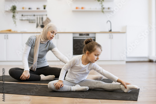 Side view of small female kid on yoga mat stretching arms and legs with slim muslim lady with closed eyes during training. Young parent and daughter in sportswear spend time happily during workout.