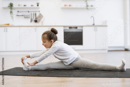Fitness, little preschool female training yoga in half tortoise pose in kitchen floor at home, copy space. Young slim girl in white sportswear makes stretching training doing splits exercise. © sofiko14