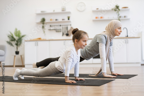 Attractive muslim mother in hijab practicing plank position with baby daughter in kitchen after wake up in morning. Feeling so comfortable and relax. Yoga of woman with her baby girl for healthcare.