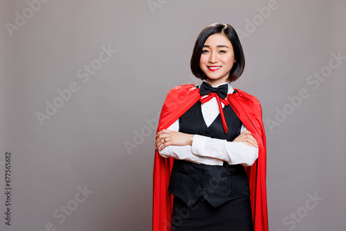 Smiling confident asian waitress in red superwoman cloak standing with arms crossed portrait. Cheerful woman receptionist with folded hands wearing hero cape looking at camera