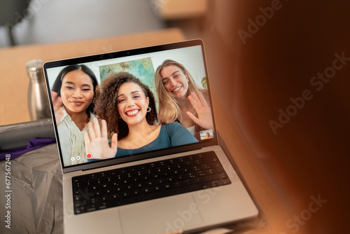Cheerful young female coworkers in casual clothes on screen of laptop waving hands and exchanging pleasantry during video call meeting with colleague in modern workspace