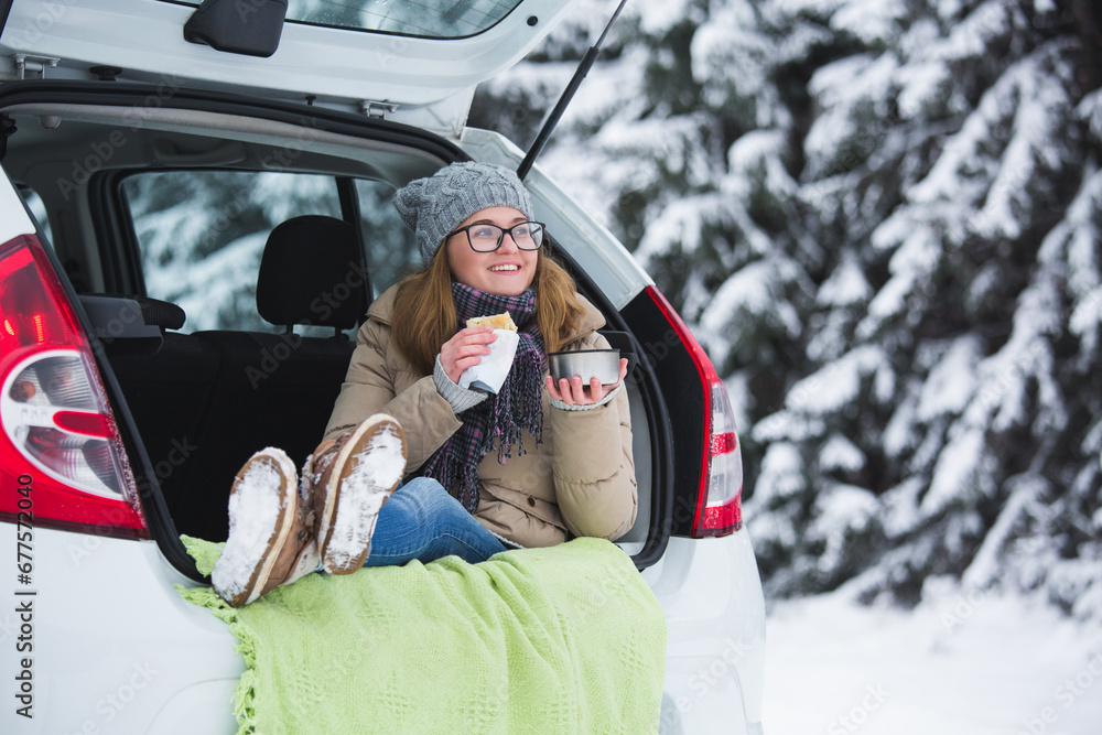 Woman in woolen hat sits in the trunk of the car and holds a cup of hot tea in her hands