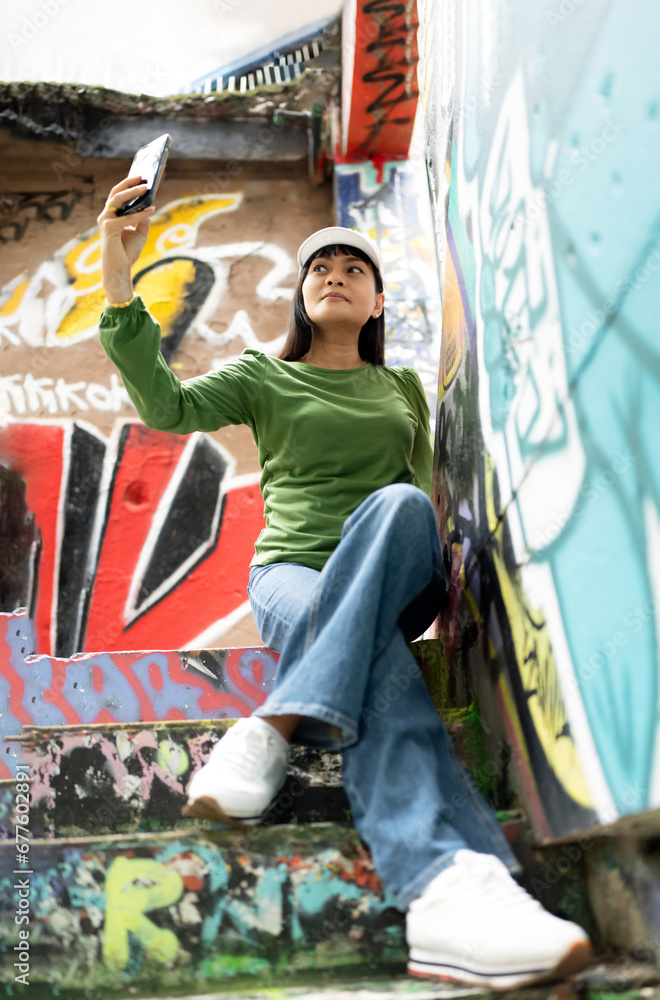 Fototapeta premium Asian female tourist in casual clothing style using mobile phone taking selfie photo while sitting on stairs on open graffiti sprayed wall art,Street Fashion Woman wearing Green Long Sleeve,Blue Jeans