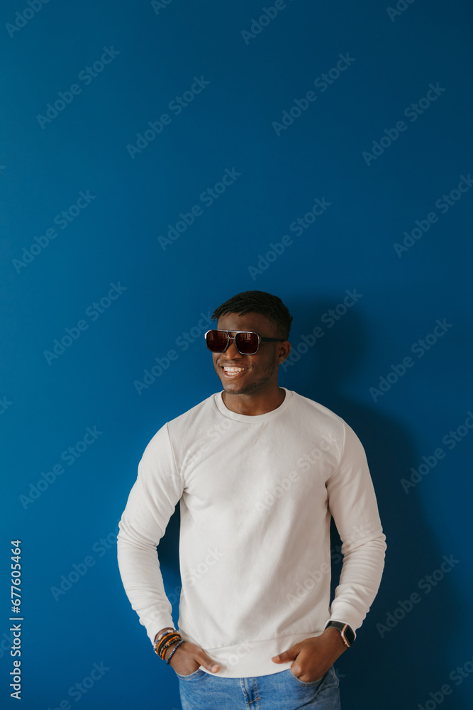 Fashionable young African man in sunglasses smiling while standing on blue background Fashionable young African man in sunglasses smiling while standing on blue background