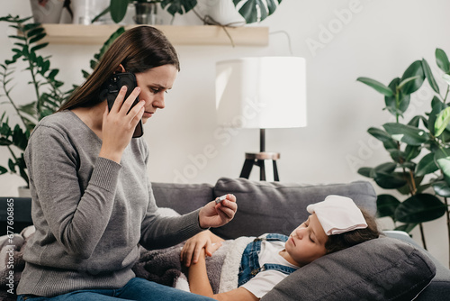 Worried young mommy calling to doctor because daughter coughing sneezing and having severe headache temperature at home. Unhealthy child with mother who distance remote talking with therapist indoors photo