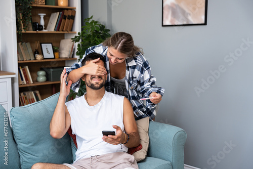 Happy wife makes surprise to husband, covers his eyes and holds pregnancy test, informs he will become father going to share good news, pose together in bedroom. Joyful future parents photo