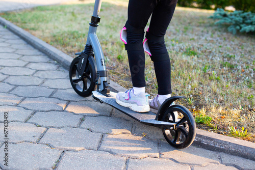 A beautiful girl in a pink protective outfit rides a scooter through the streets of the city in summer. Safe cycling and scootering without injury.