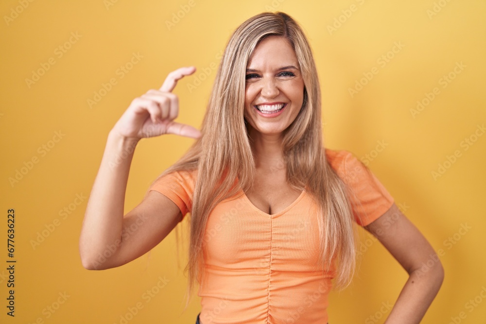 Young woman standing over yellow background smiling and confident gesturing with hand doing small size sign with fingers looking and the camera. measure concept. Young woman standing over yellow background smiling and confident gesturing with hand doing small size sign with fingers looking and the camera. measure concept.