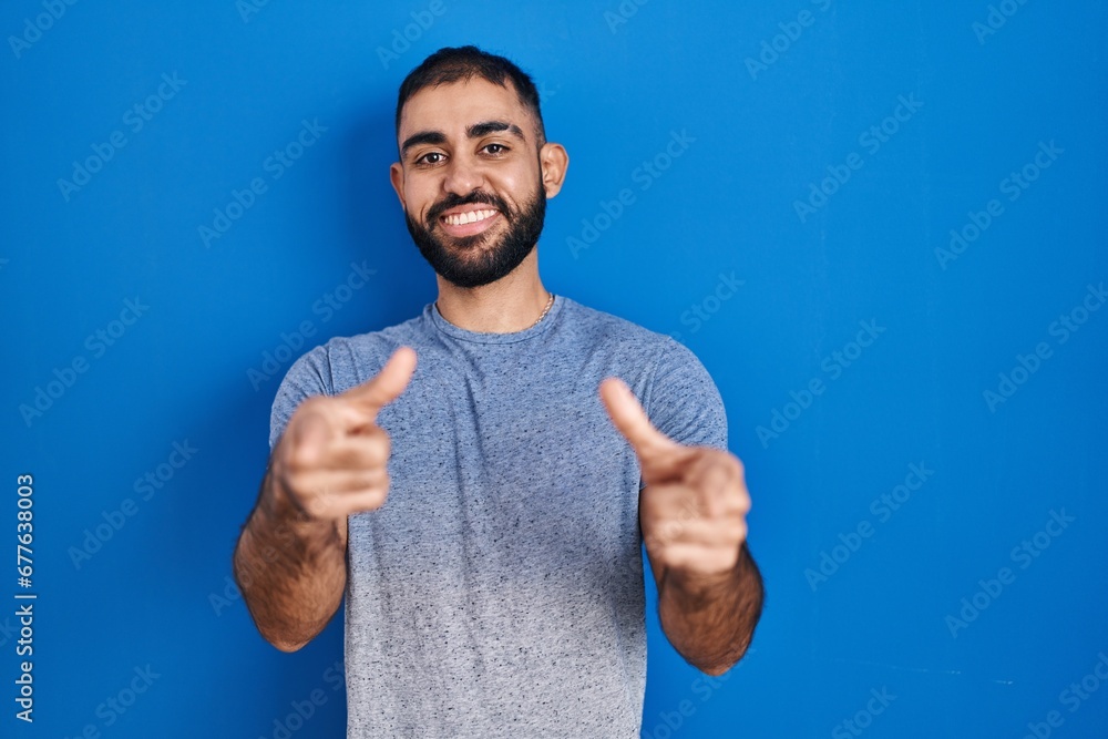 Middle east man with beard standing over blue background pointing fingers to camera with happy and funny face. good energy and vibes. Middle east man with beard standing over blue background pointing fingers to camera with happy and funny face. good energy and vibes.