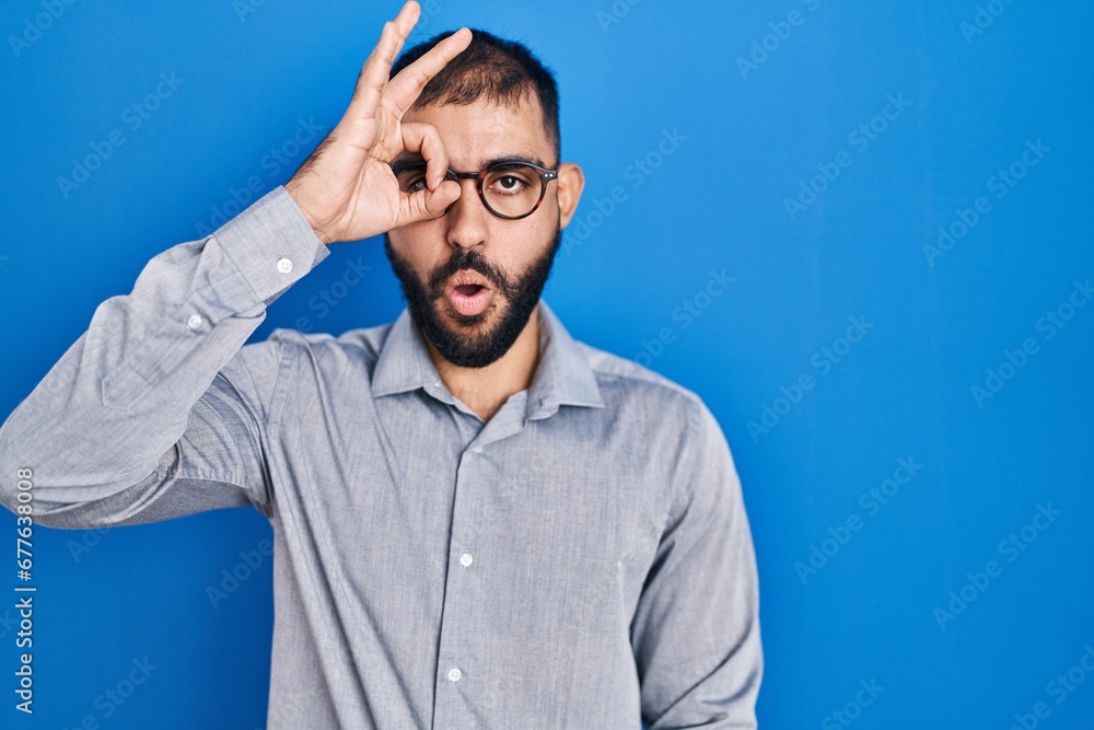 Middle east man with beard standing over blue background doing ok gesture shocked with surprised face, eye looking through fingers. unbelieving expression.
