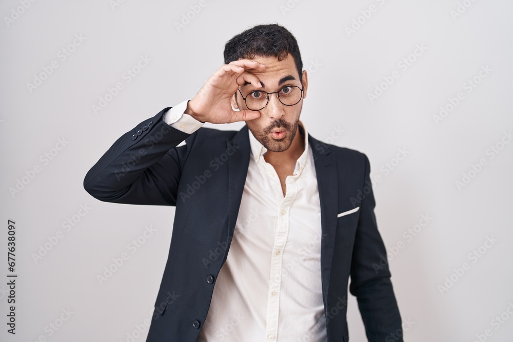 Handsome business hispanic man standing over white background doing ok gesture shocked with surprised face, eye looking through fingers. unbelieving expression. Handsome business hispanic man standing over white background doing ok gesture shocked with surprised face, eye looking through fingers. unbelieving expression.