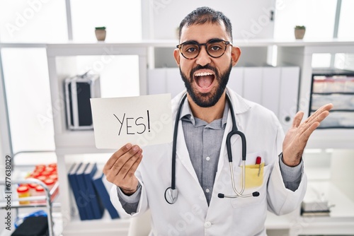 Middle east man with beard wearing doctor uniform and stethoscope holding yes banner celebrating achievement with happy smile and winner expression with raised hand