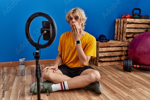 Young man recording training tutorial at the gym covering mouth with hand, shocked and afraid for mistake. surprised expression