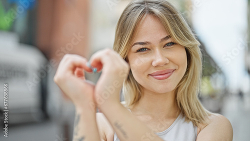 Young blonde woman smiling confident doing heart gesture with hands at street