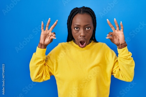 Beautiful black woman standing over blue background looking surprised and shocked doing ok approval symbol with fingers. crazy expression