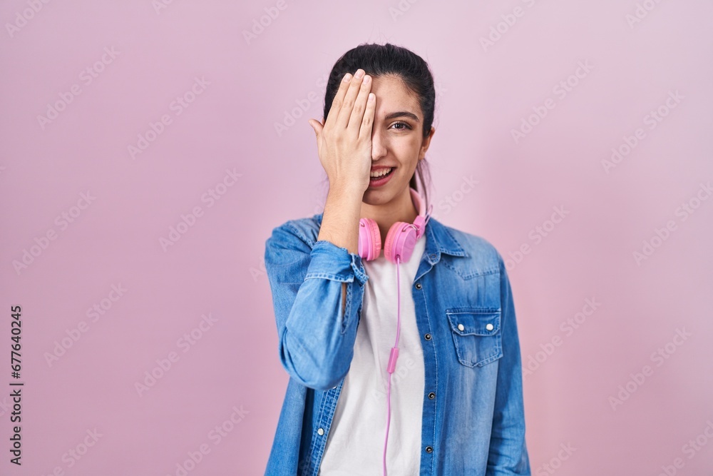 Young beautiful woman standing over pink background covering one eye with hand, confident smile on face and surprise emotion. Young beautiful woman standing over pink background covering one eye with hand, confident smile on face and surprise emotion.