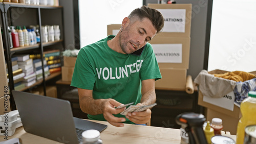 Handsome young hispanic man volunteering, counting donation dollars while engaged in a heartfelt conversation on his smartphone at the charitable center