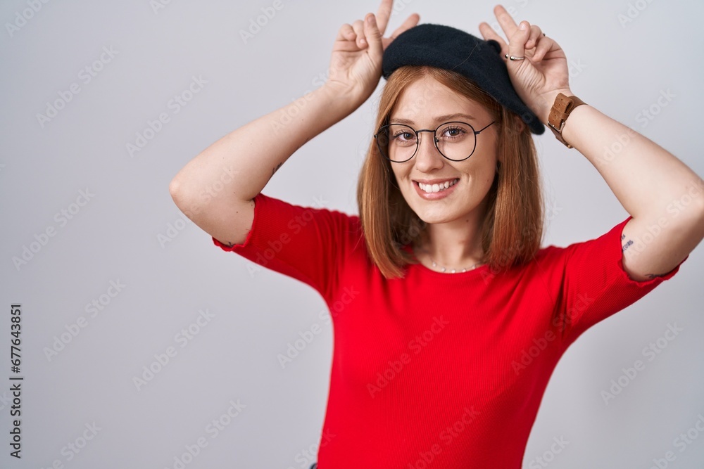 Young redhead woman standing wearing glasses and beret posing funny and crazy with fingers on head as bunny ears, smiling cheerful