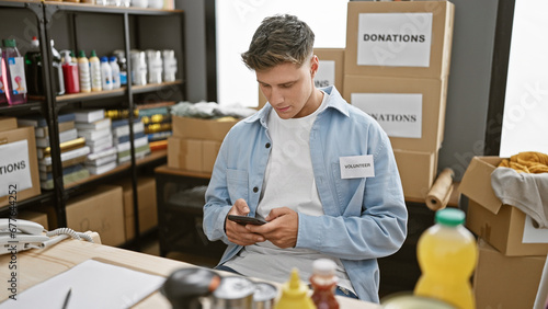 Handsome bearded young caucasian man volunteering at a local charity center, focused on his smartphone as he types out messages for donations, sitting indoors, surrounded by cardboard boxes