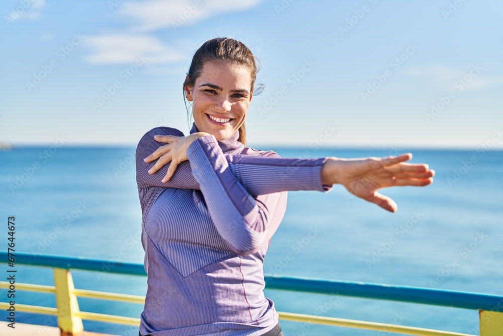 Young beautiful hispanic woman wearing sportswear stretching arm at seaside Young beautiful hispanic woman wearing sportswear stretching arm at seaside